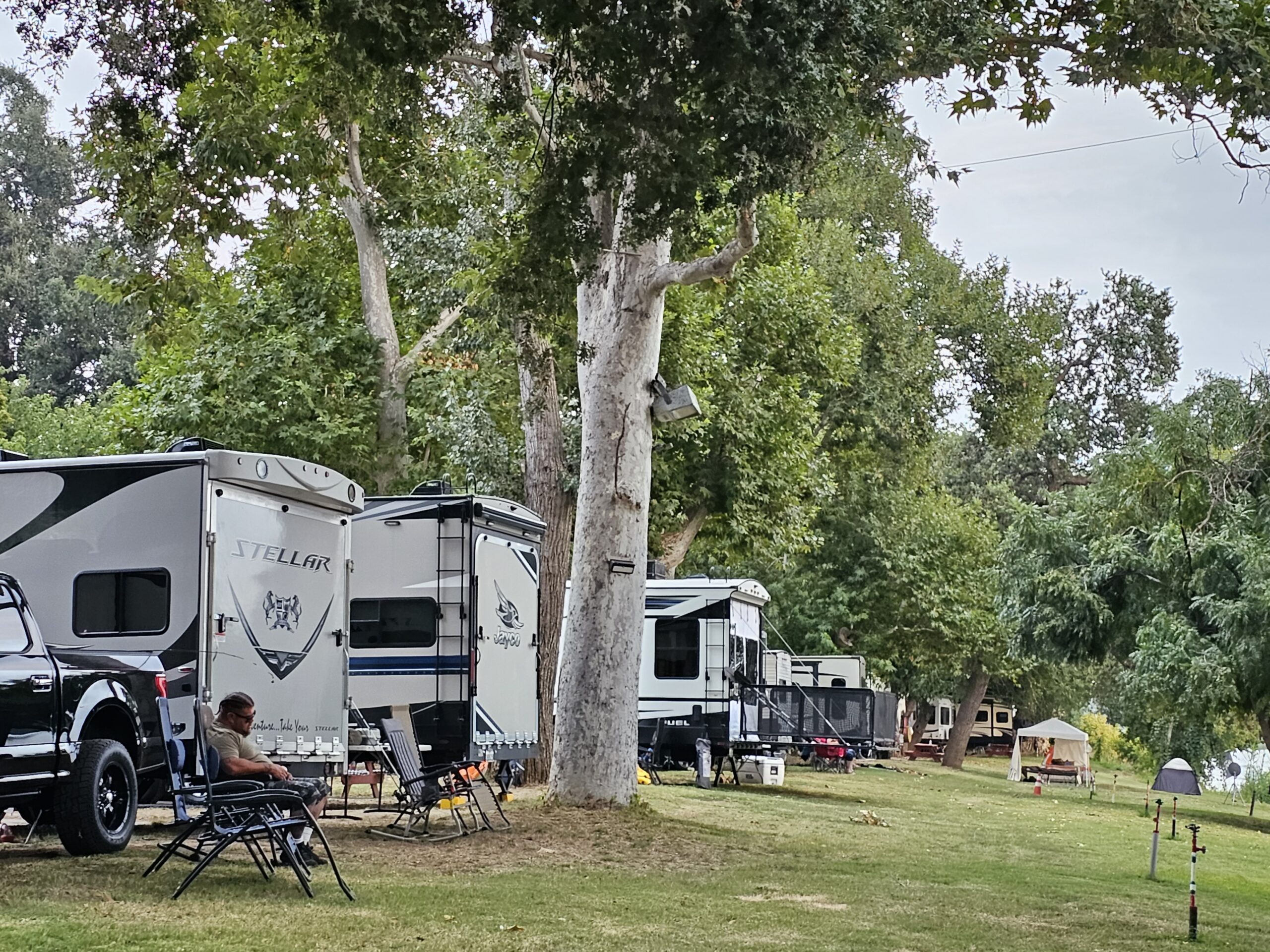 Several RVs and campers are parked on grassy land under tall trees. A person sits in a chair near one RV, and camping chairs and tables are set up nearby. The setting appears peaceful and outdoorsy.