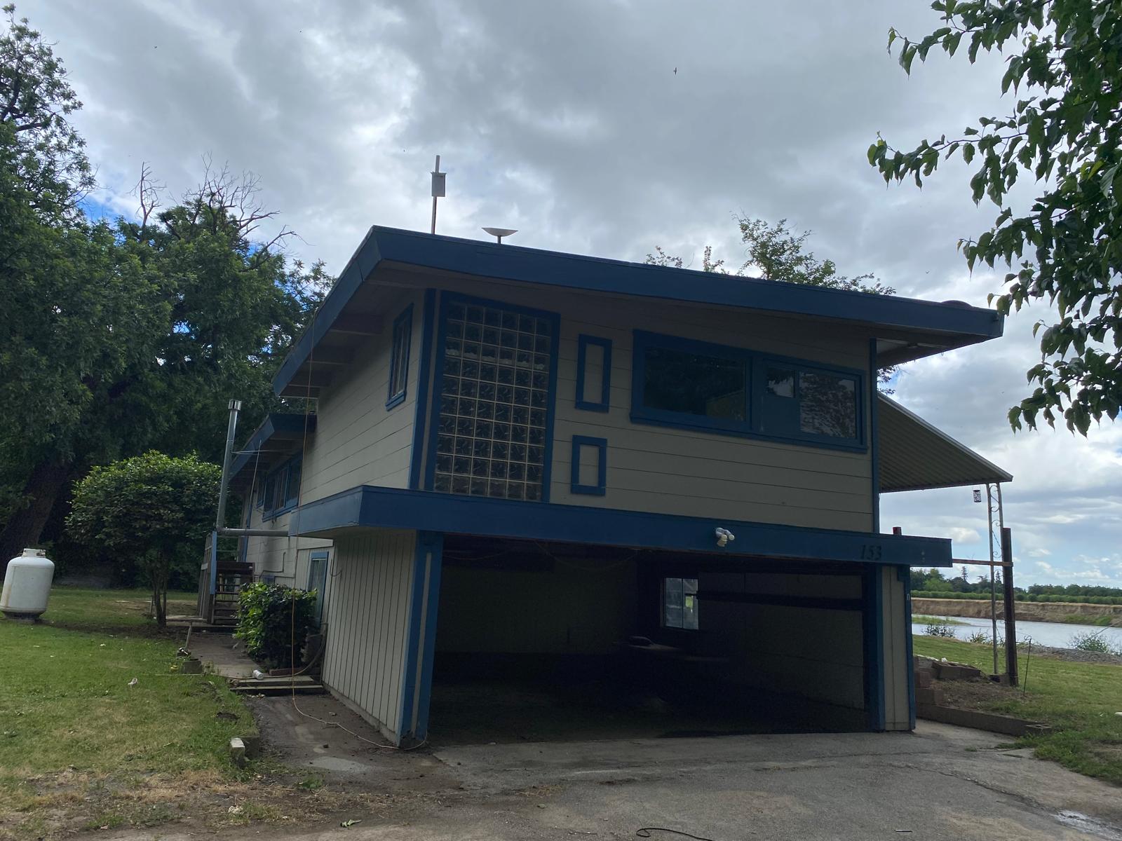 Two-story house with a blue and beige exterior, glass block window, and carport underneath, situated near a body of water with cloudy skies overhead.