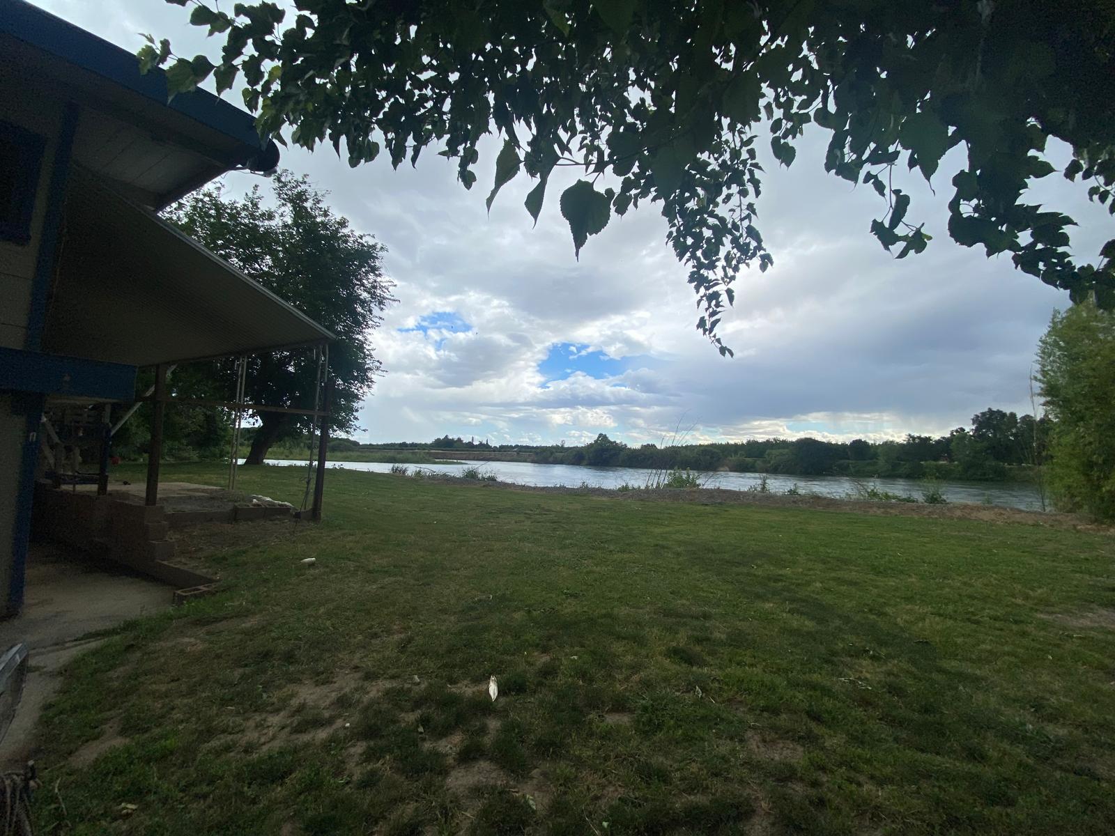 A grassy yard beside a house overlooks a calm river, with trees and cloudy sky in the background.
