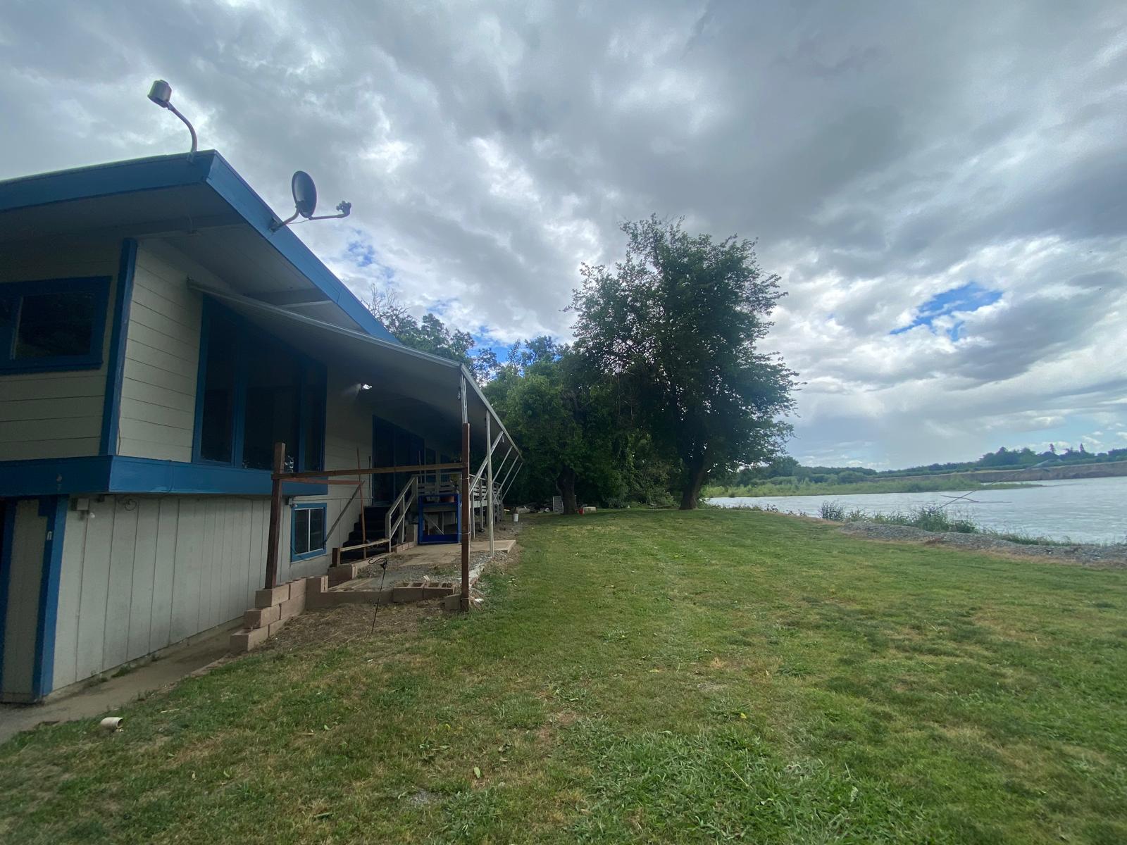 A house with blue trim sits beside a grassy lawn near a body of water under a cloudy sky.