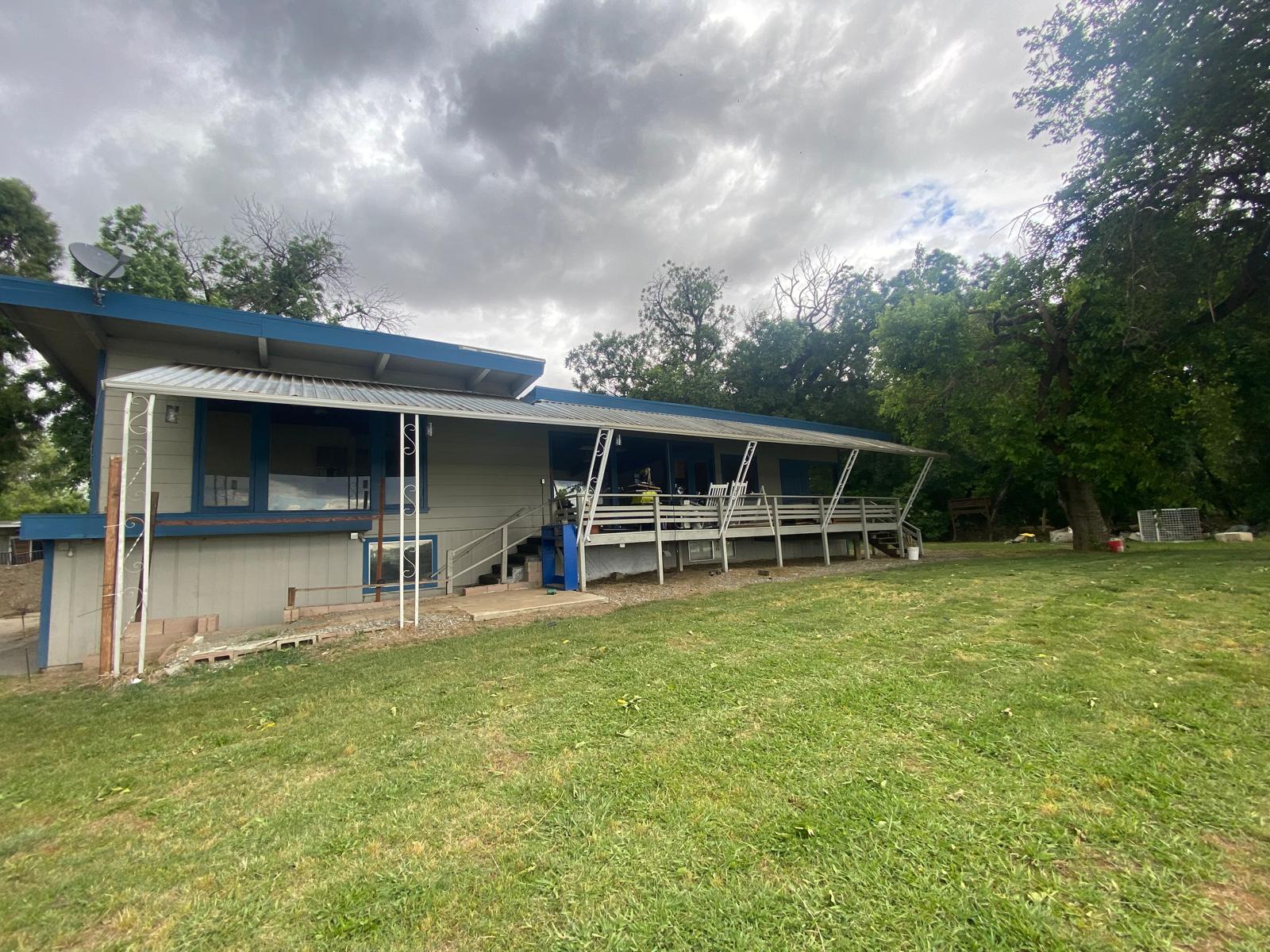 Single-story building with blue trim, covered porch, and metal supports, surrounded by grass and trees under a cloudy sky.