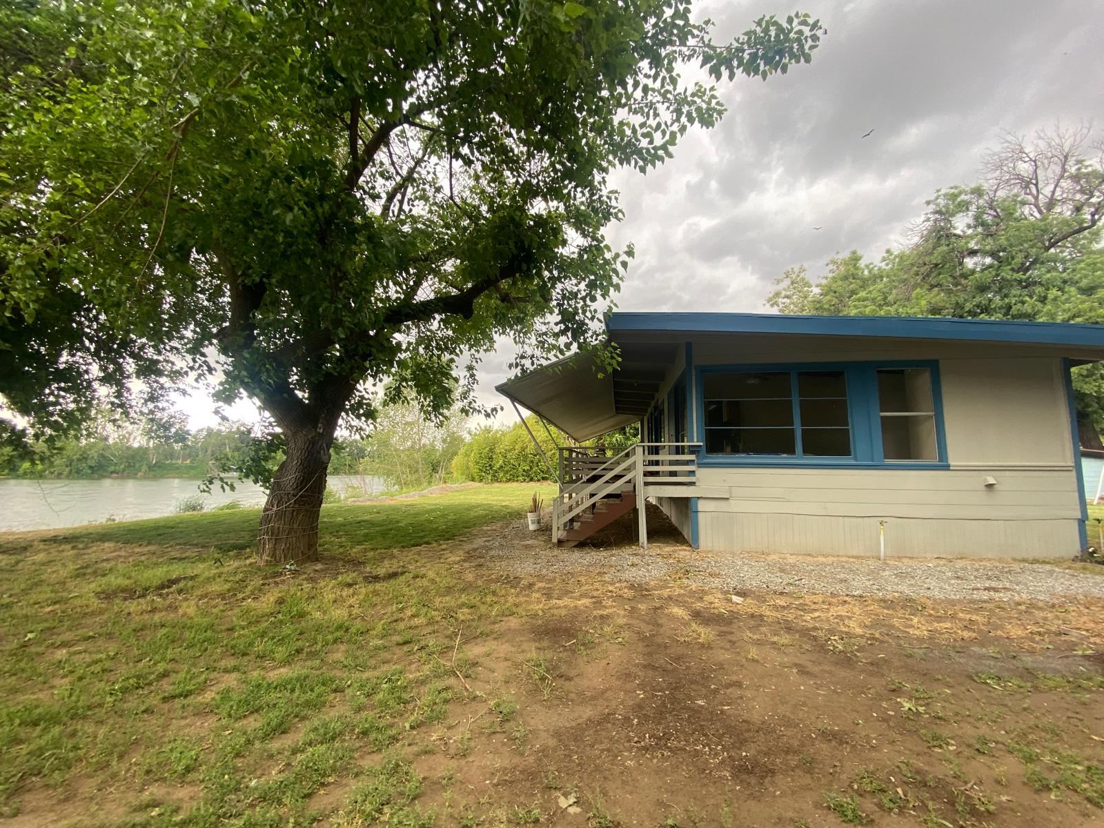 A single-story house with blue trim and a covered porch, next to a large tree, sits beside a grassy yard and a body of water under a cloudy sky.