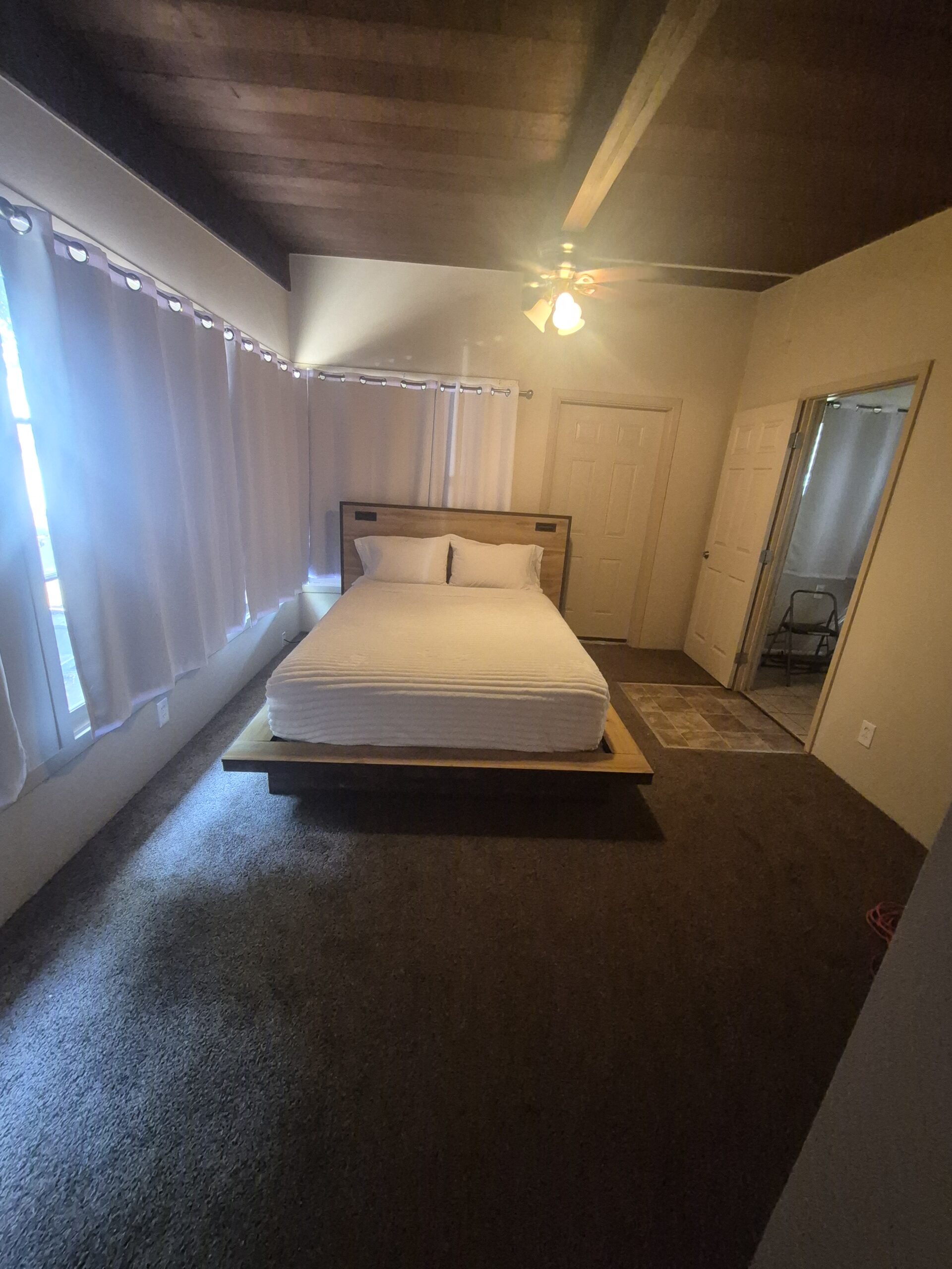 A bedroom with a bed centered against the wall, white bedding, brown carpet, curtained windows, ceiling fan, and a door leading to another room.