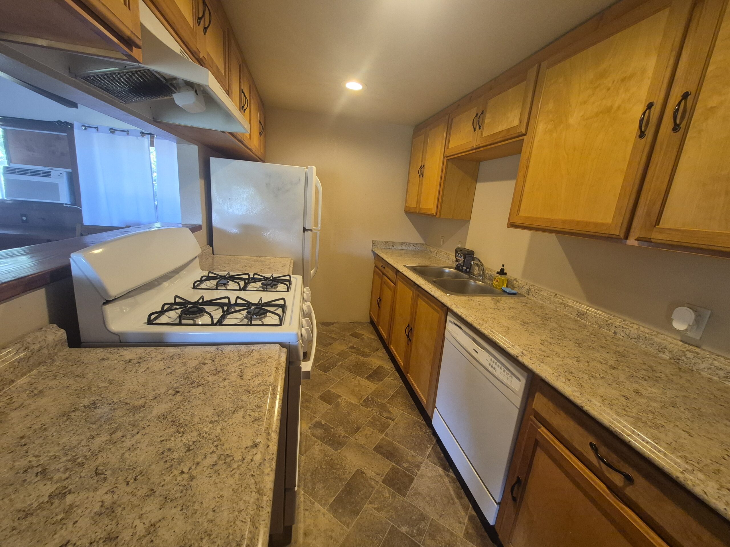 Narrow kitchen with wood cabinets, a gas stove, white refrigerator, dishwasher, sink, and granite-style countertops under warm lighting.