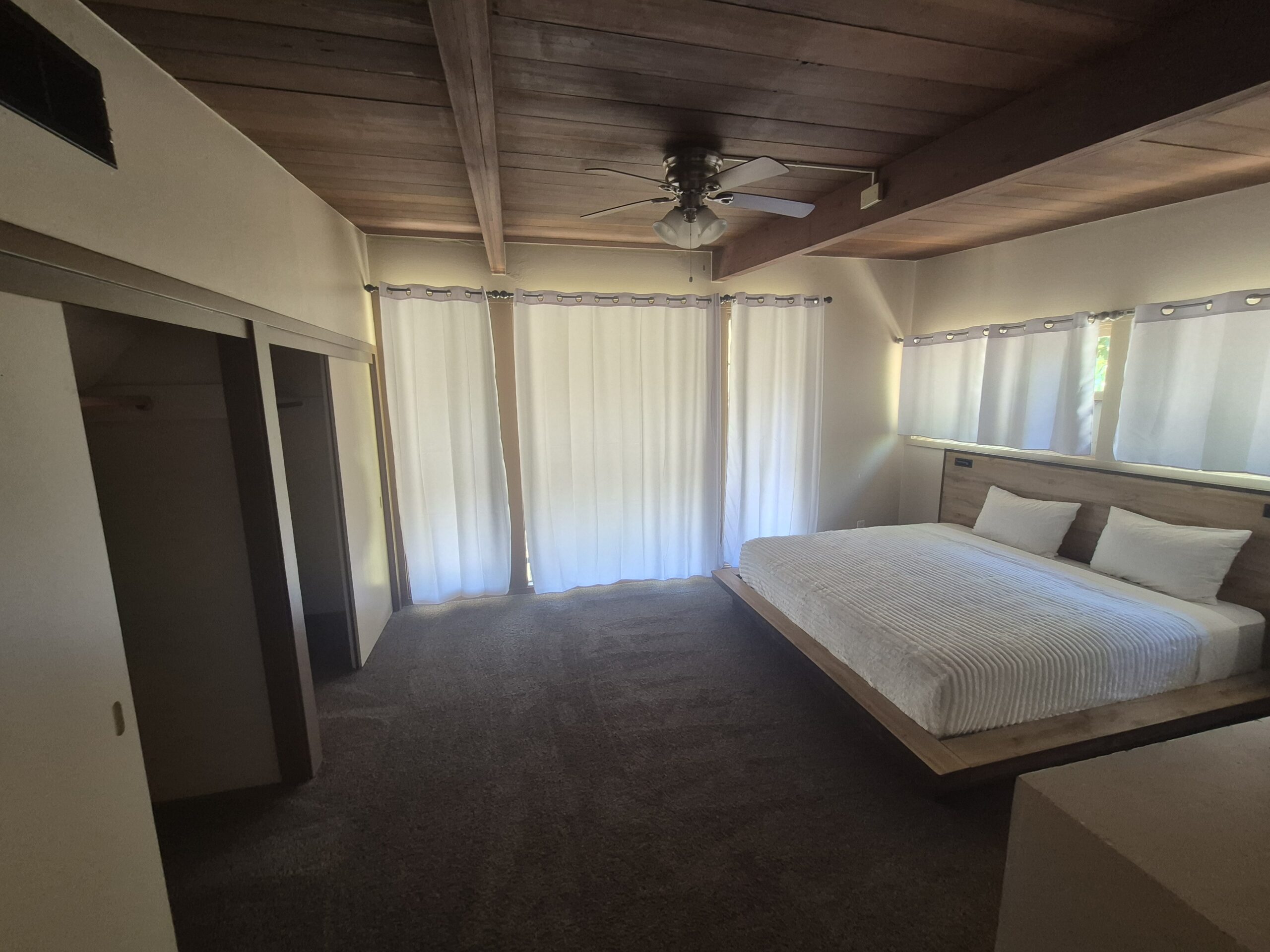 A bedroom with a large bed, three pillows, carpeted floor, ceiling fan, and closed curtains covering windows and a sliding glass door.