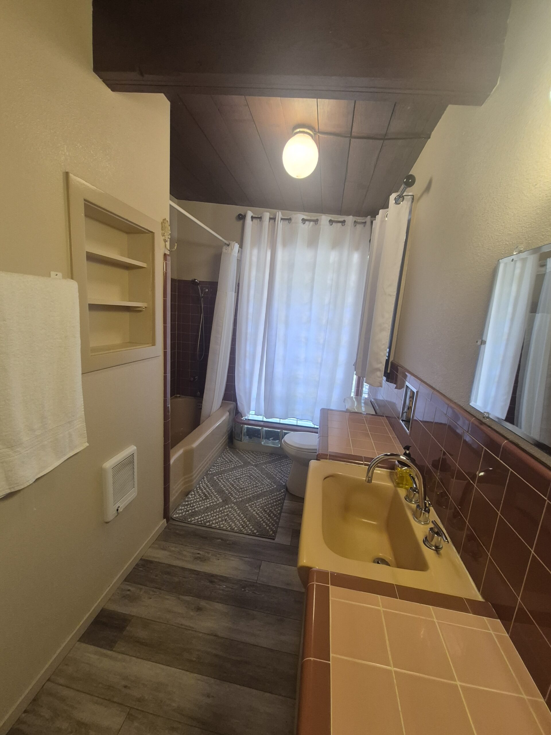 Bathroom with yellow sink, brown-tiled countertop, brown bathtub and shower, toilet, large mirror, towel rack, and wood ceiling under warm light.