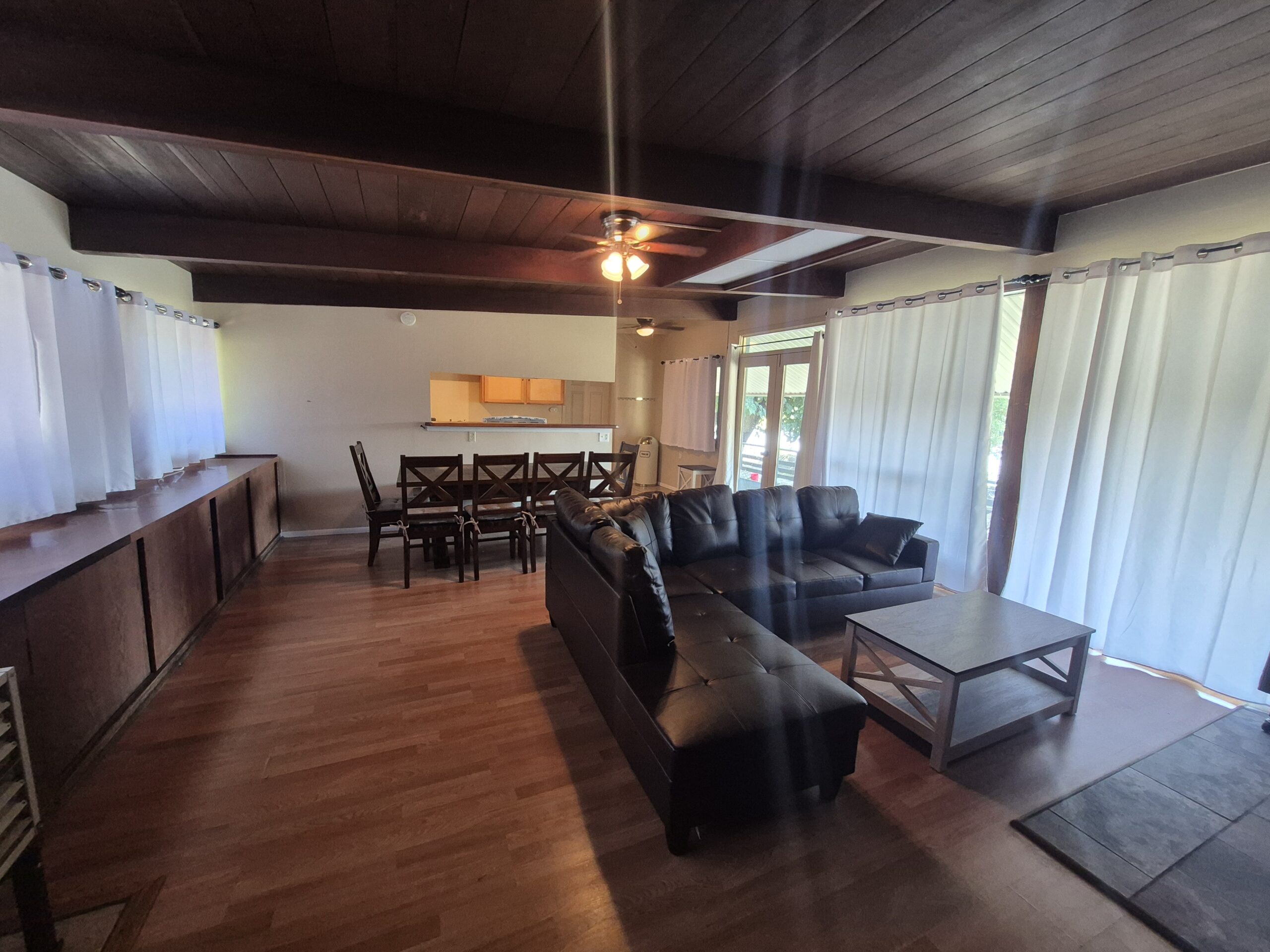 Open living and dining area with wooden floors, dark wood ceiling beams, a black sectional sofa, dining table with chairs, and large windows covered by white curtains.