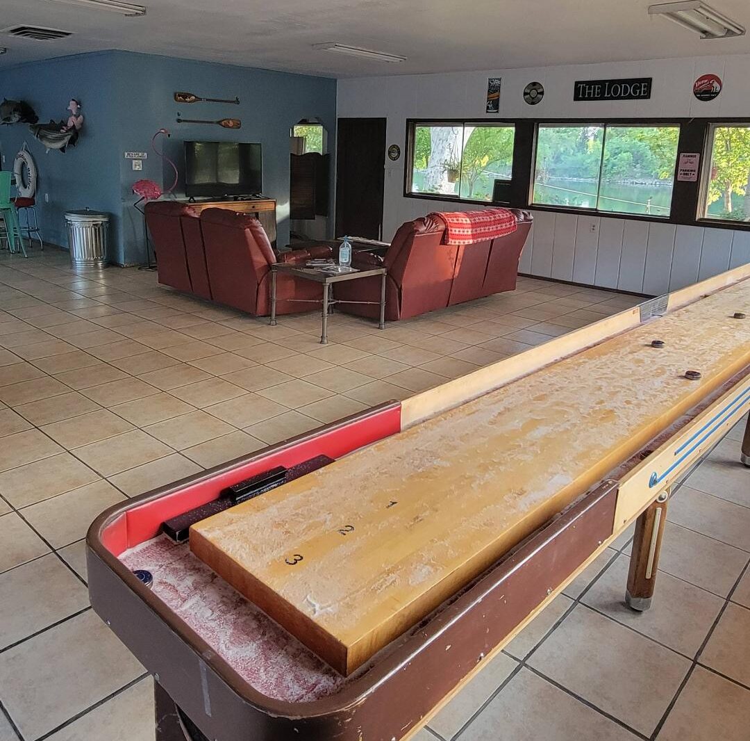 A lounge area with red couches, a TV, and wall decor, featuring a shuffleboard table in the foreground on a tiled floor.